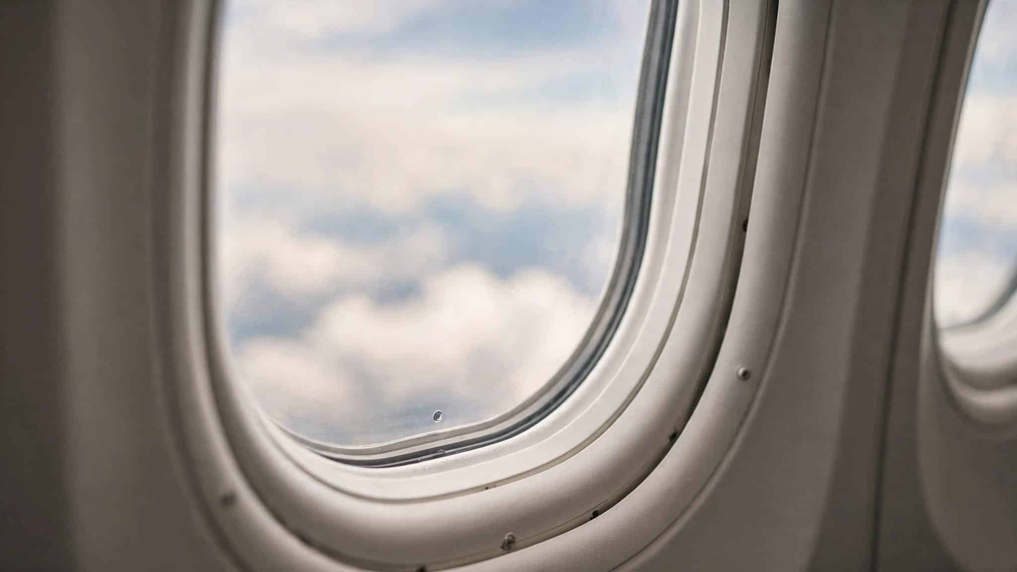 A tiny hole sits near the bottom of an airplane window, with clouds beyond the glass.